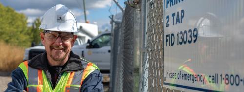 Man wearing a hard hat, safety glasses and a safety vest smiles at the camera outside a FortisBC station